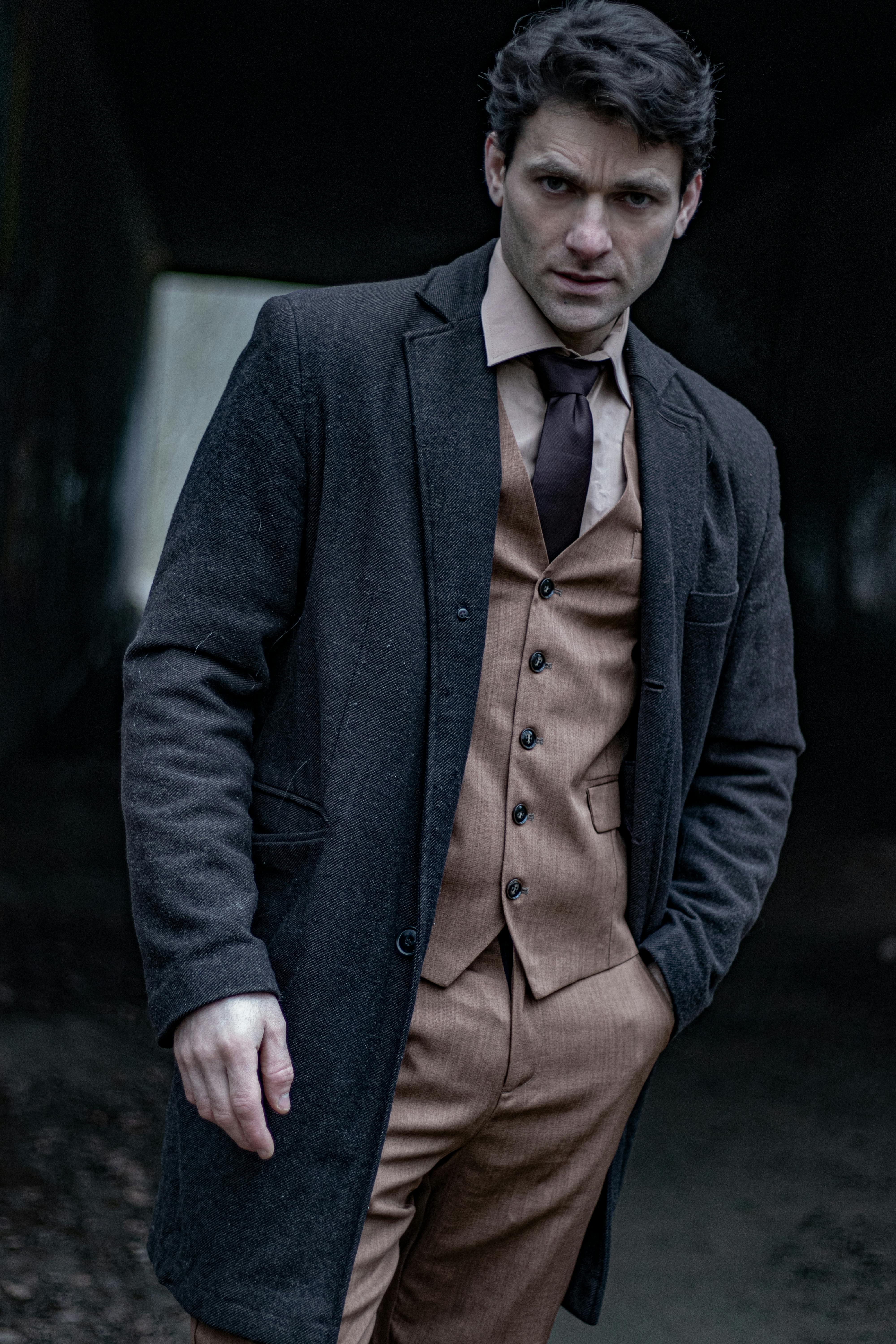 A man wearing a vintage suit and overcoat poses stylishly indoors with a dramatic expression.
