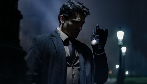 Free A man in dark clothing holds a pocket watch on a dimly lit street at night. Stock Photo