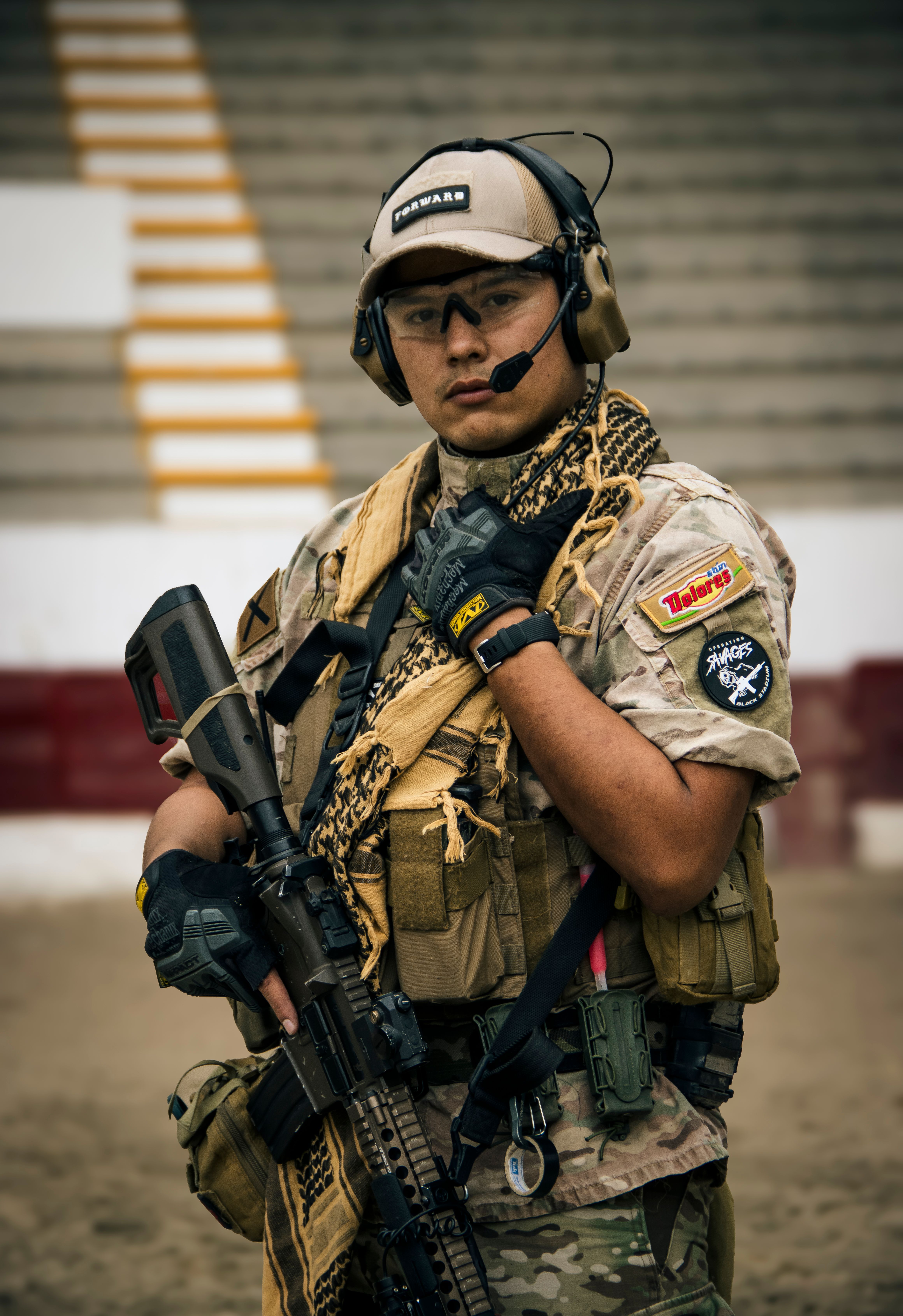 A soldier in tactical gear with rifle and headset standing in an outdoor arena.