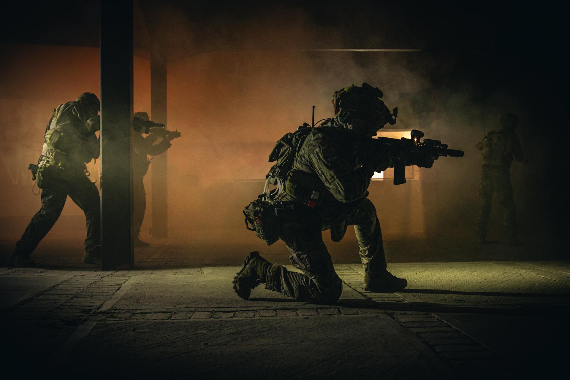 Four soldiers in tactical gear engage in a smoky nighttime military operation indoors.