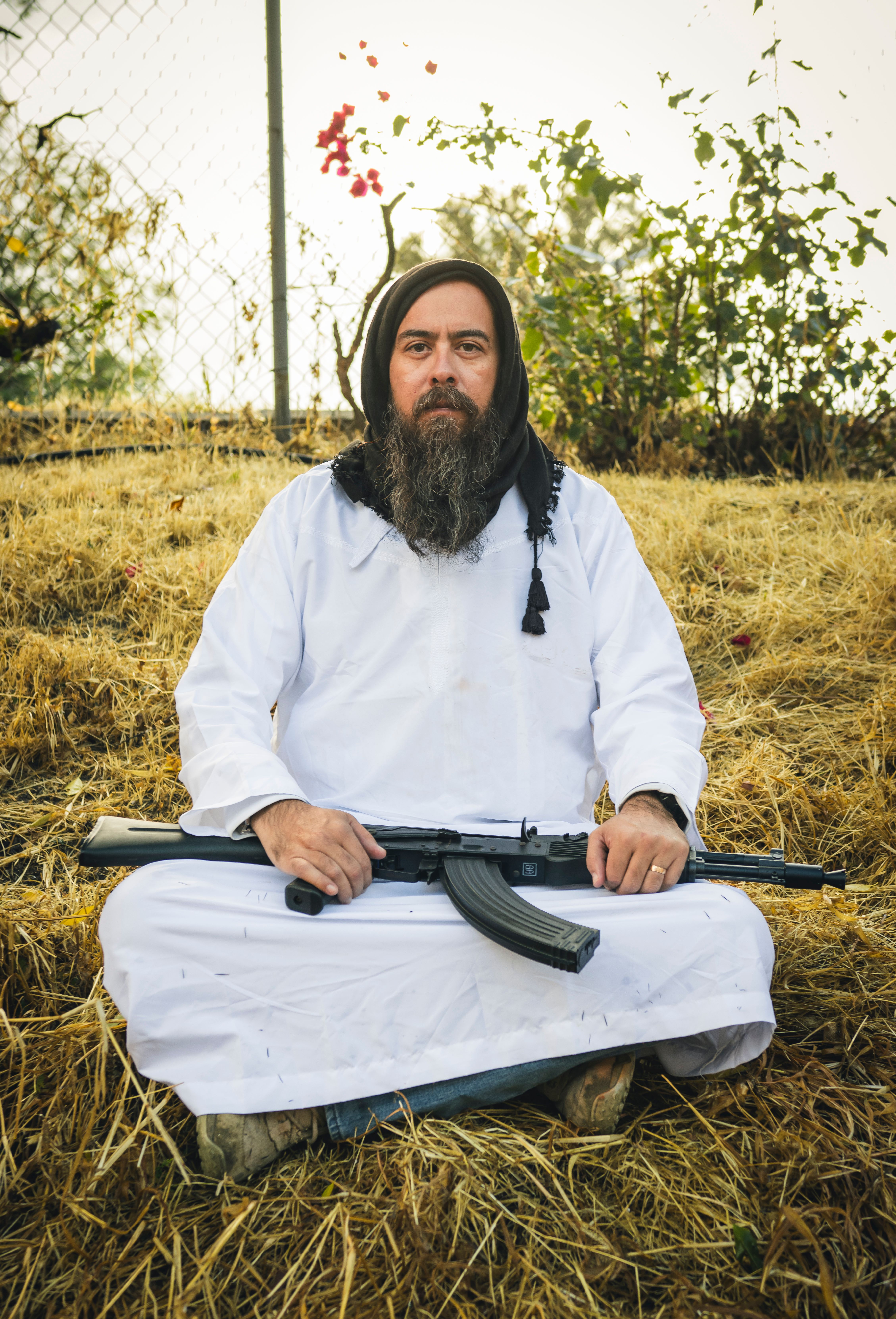 Sitting man in white traditional attire holding rifle in grassy outdoor setting.