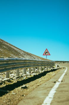 A winding mountain road under a bright blue sky, featuring a warning sign.