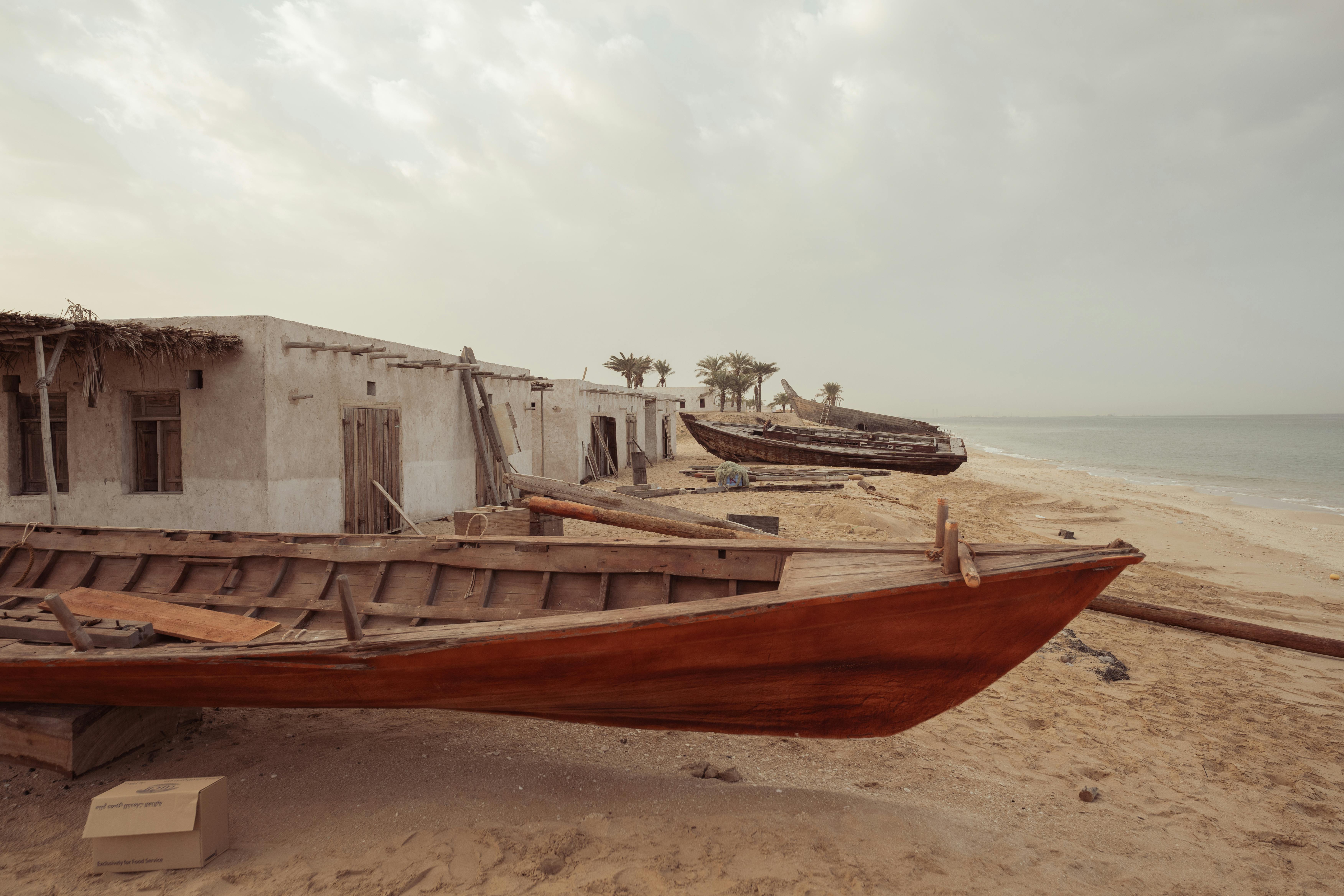 Rustic wooden boats lining an empty sandy beach beside old stone houses under a cloudy sky.