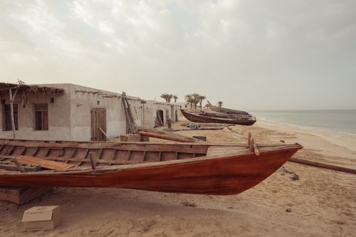 Free Rustic wooden boats lining an empty sandy beach beside old stone houses under a cloudy sky. Stock Photo