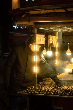 A vendor roasts chestnuts at a street market stall, illuminated by warm lights.