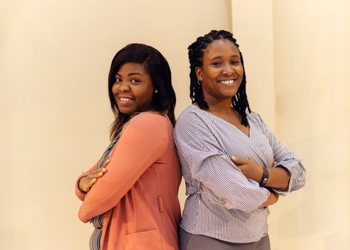 Two women standing back to back, smiling confidently indoors, symbolizing unity and empowerment.