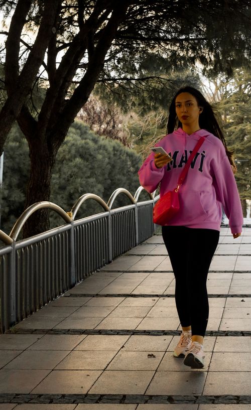 Free Woman in pink hoodie walking outdoors with smartphone in hand. Stock Photo