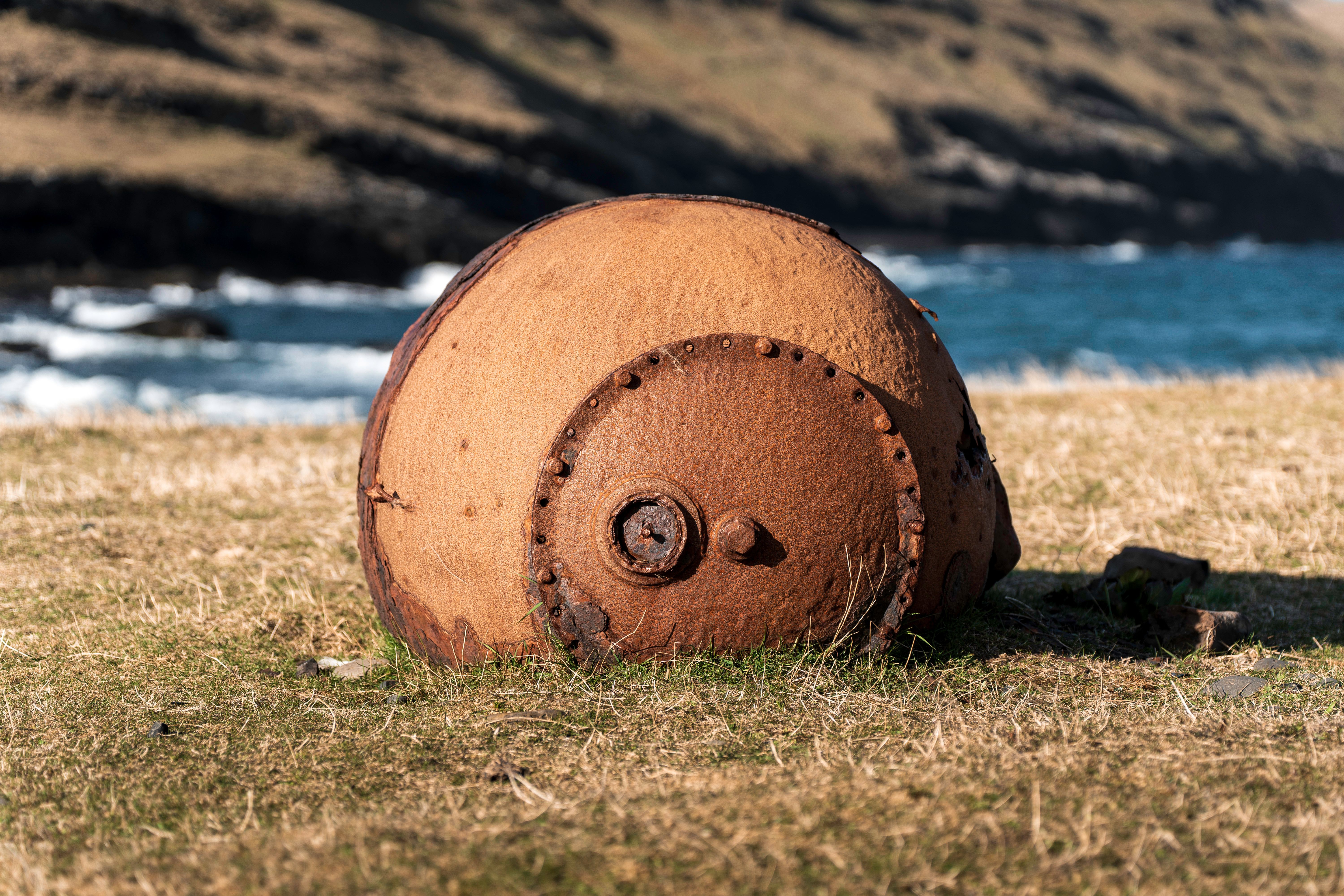 A large, corroded metal object lying on a grassy area near the water.