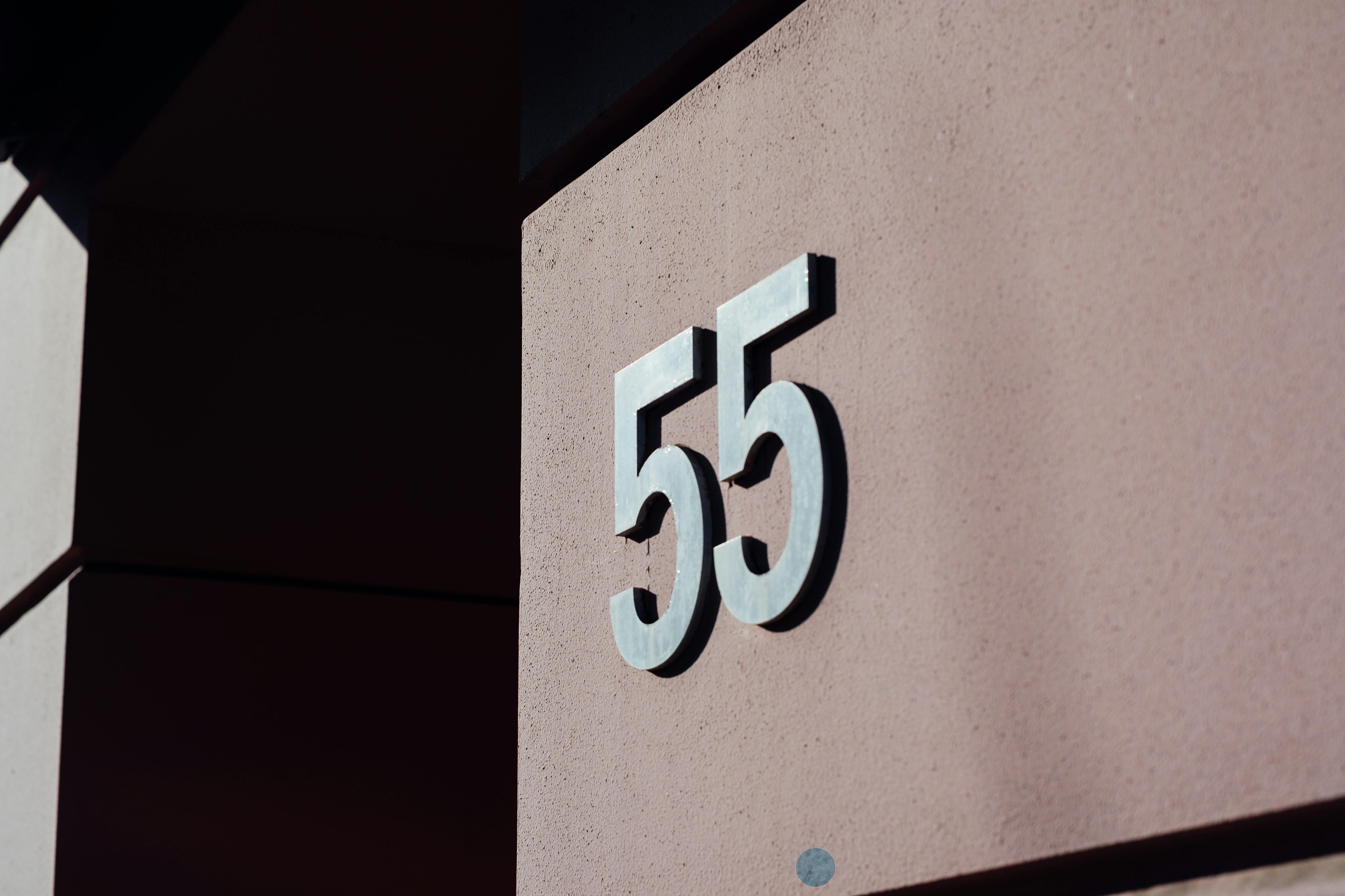 Close-up of a modern building wall with metal number 55 casting a shadow.