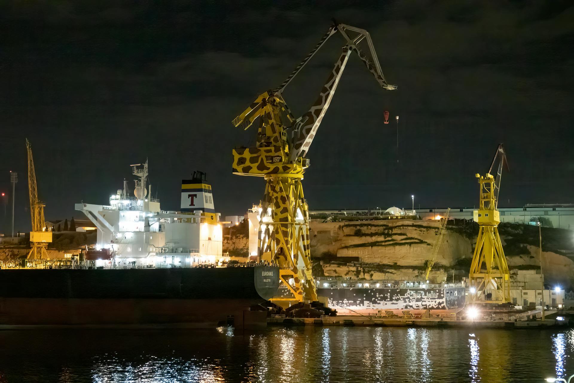 A well-lit shipyard in Malta featuring large cranes and a cargo ship under the night sky.