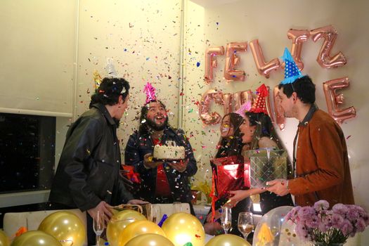 Group celebrating a birthday with cake, balloons, and confetti indoors.