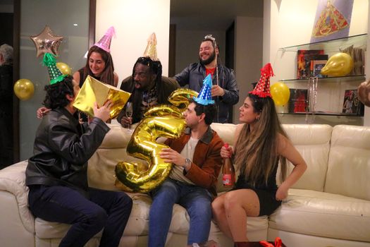 Group of friends celebrating a birthday with party hats and balloons indoors.