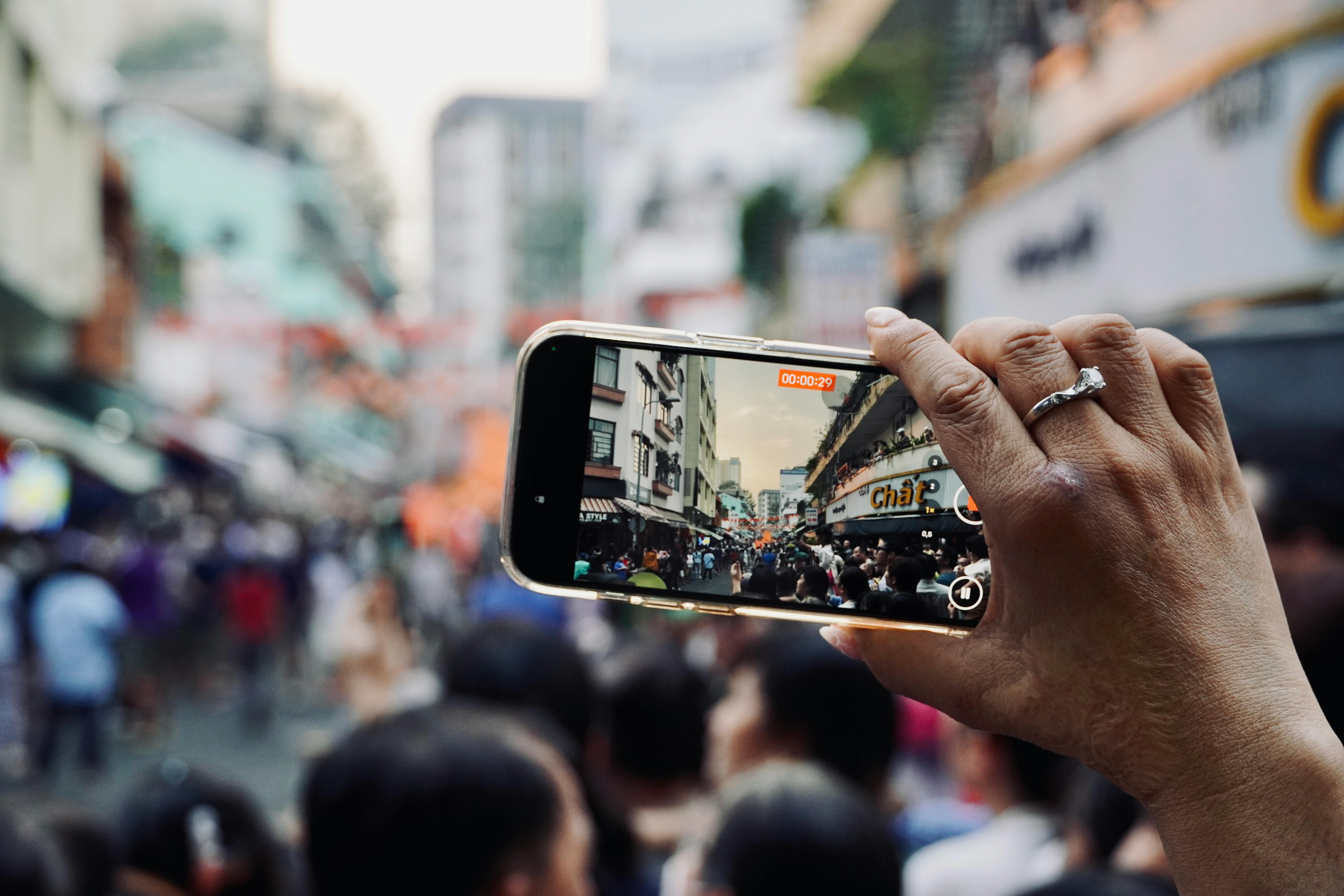 Free Hand holding smartphone capturing bustling city street scene with crowd. Stock Photo