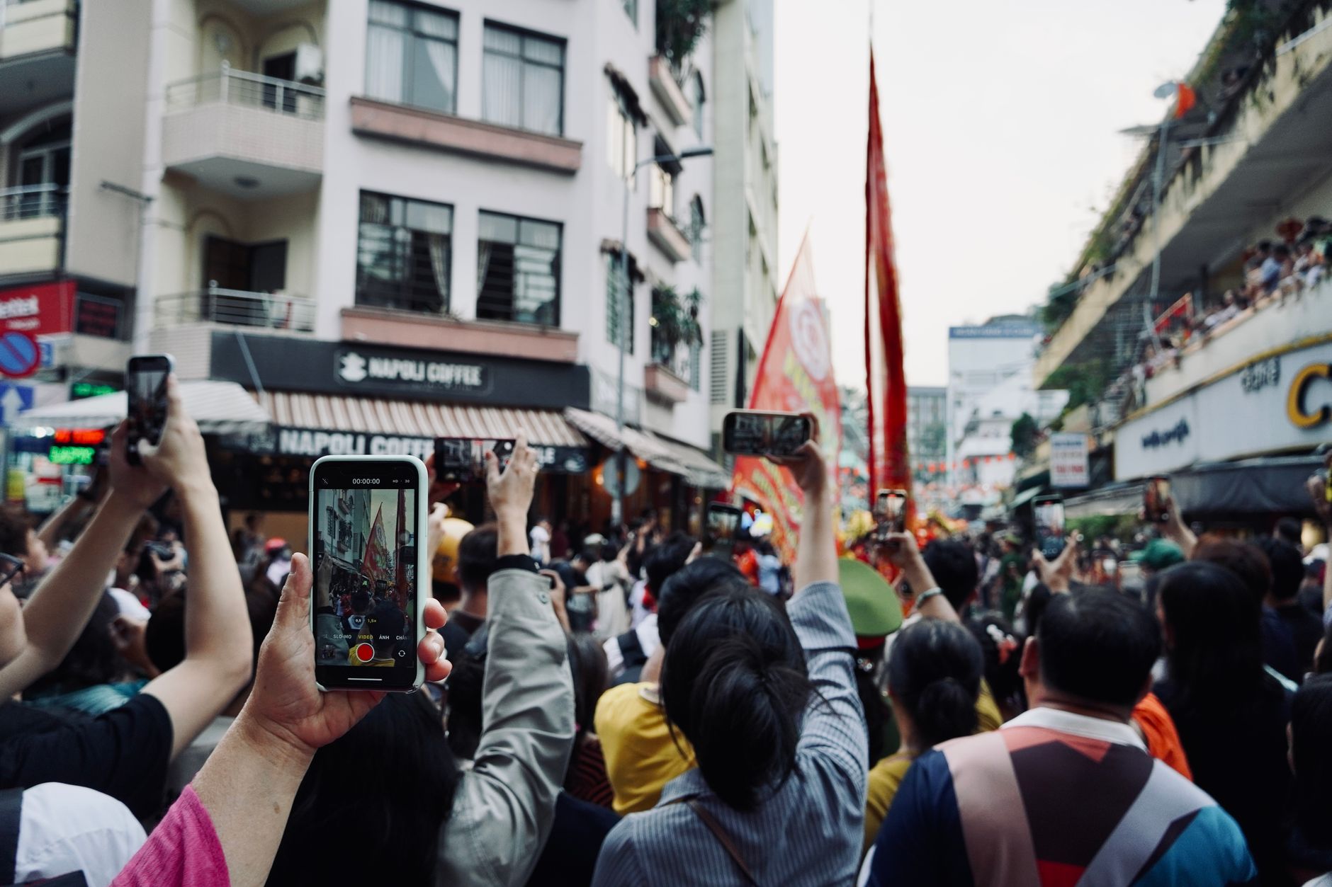 A vibrant street parade captured by many people using smartphones in a bustling urban scene.