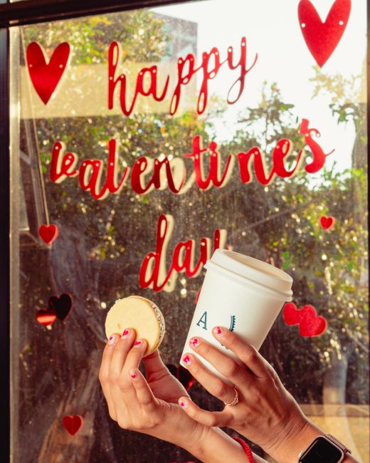 Hands holding coffee and macaron in front of Valentine's Day window display.