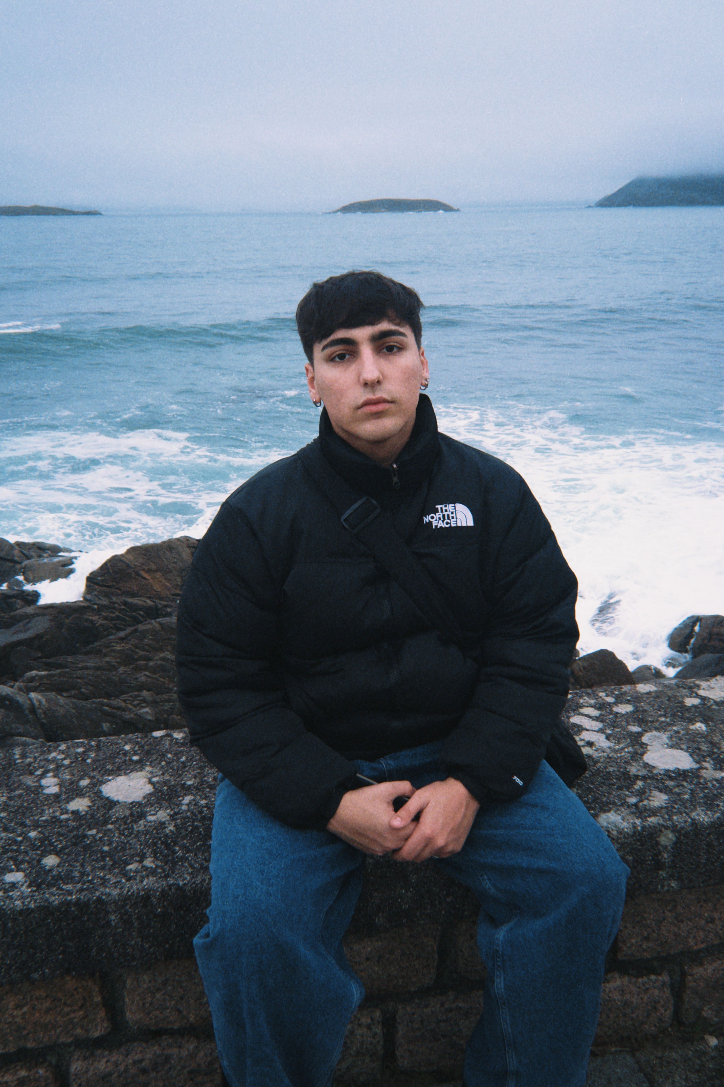 A young man sits on a stone wall with the ocean in the background, wearing a black jacket.