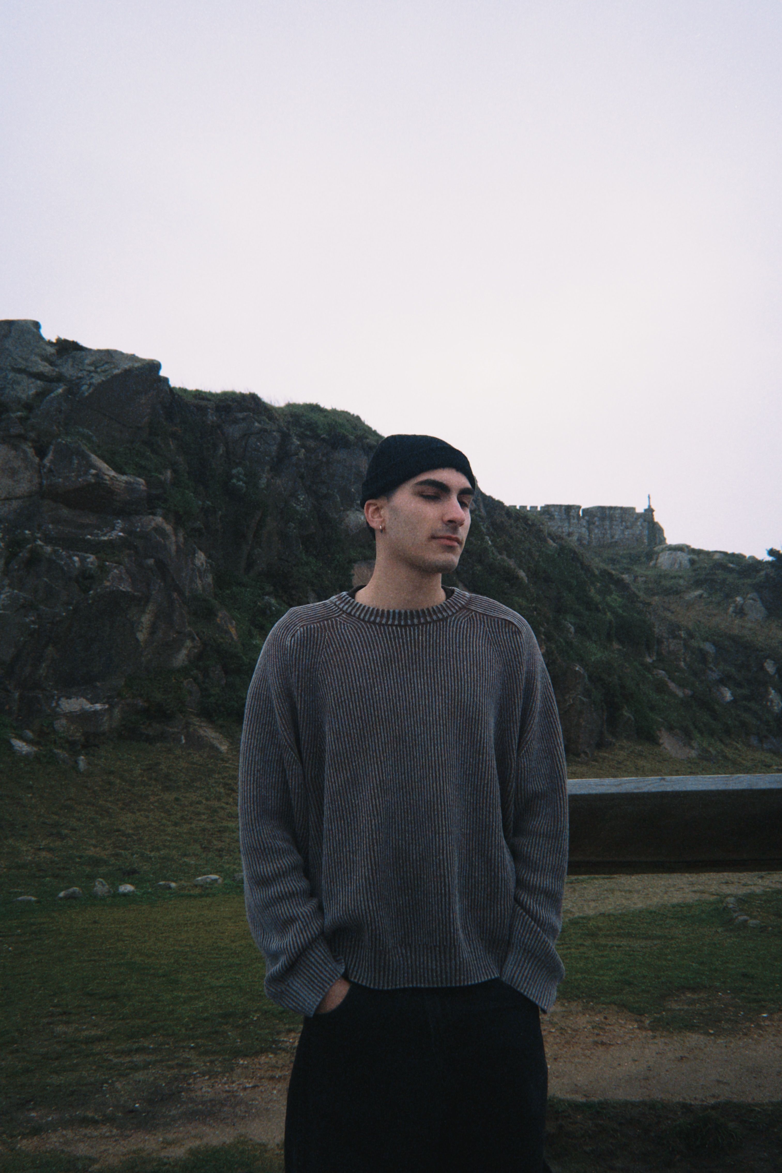 Young man in a sweater stands outdoors against a rocky landscape during daytime.