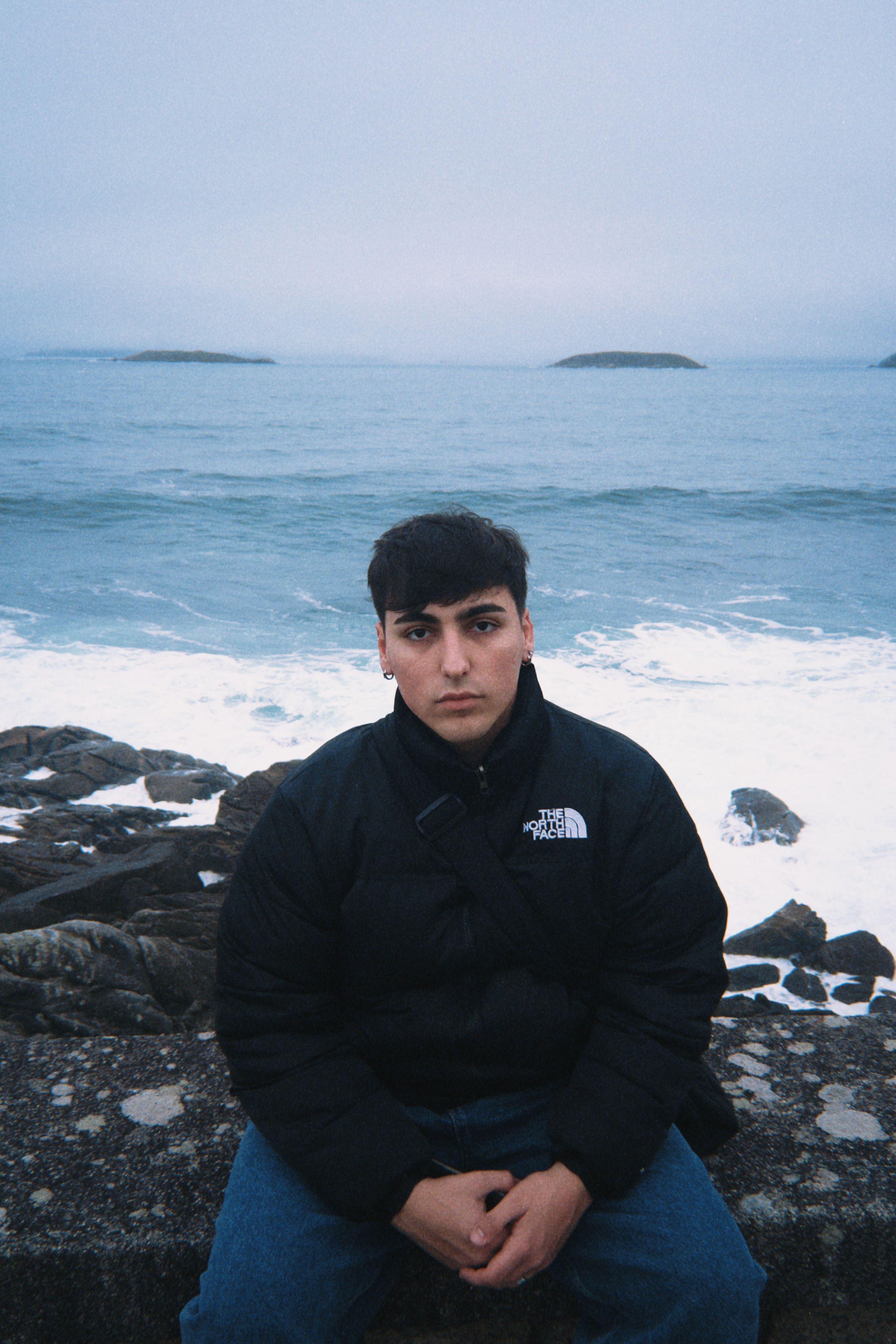 Man in a black jacket sitting by the ocean with rocky shore in the background.