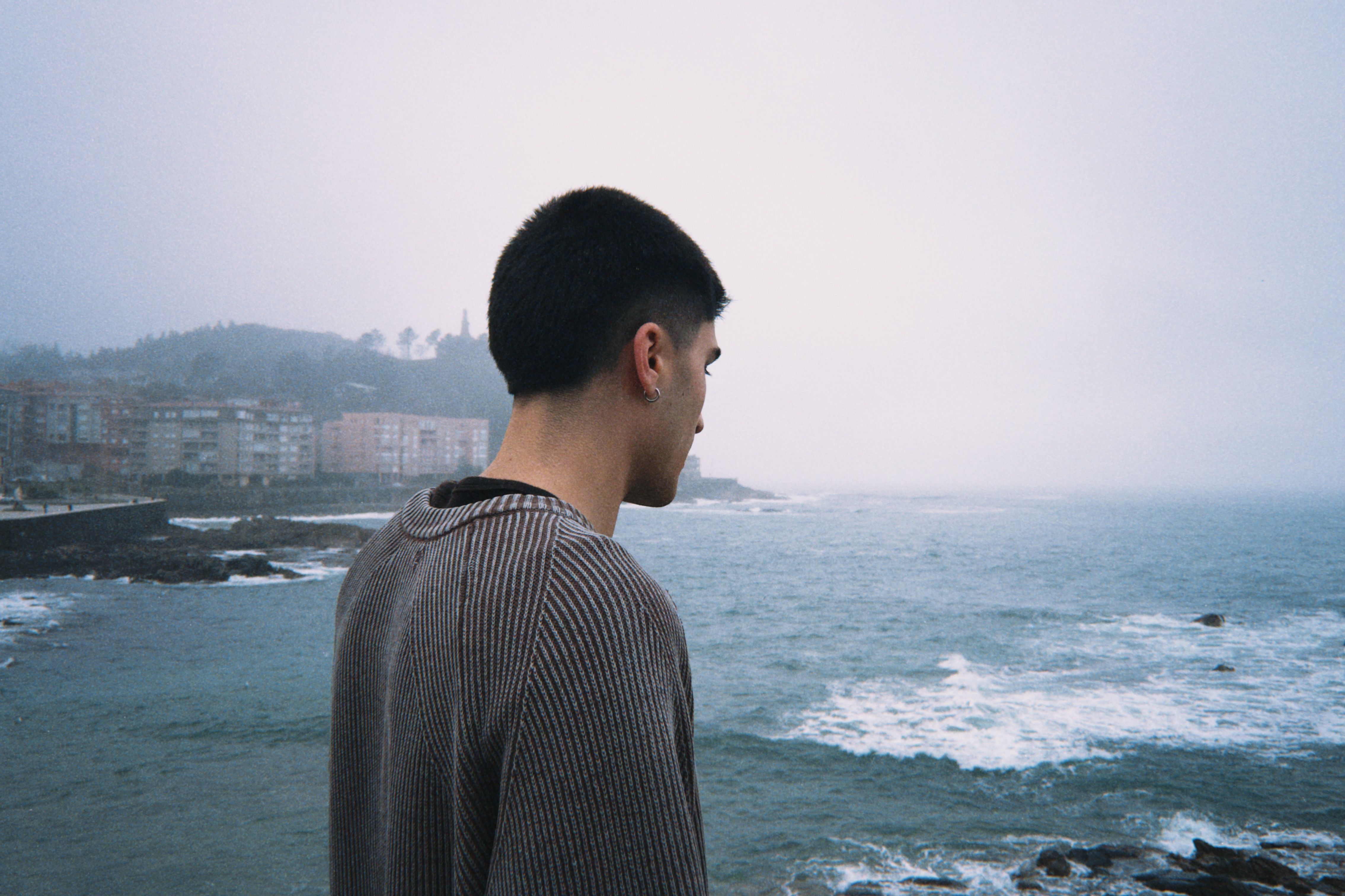 A young man stands by the ocean, gazing thoughtfully at the foggy, wave-filled landscape.