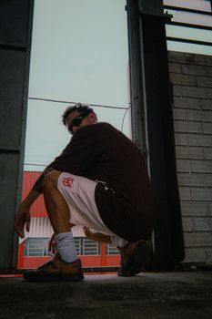Man squatting stylishly inside a warehouse with an industrial backdrop.
