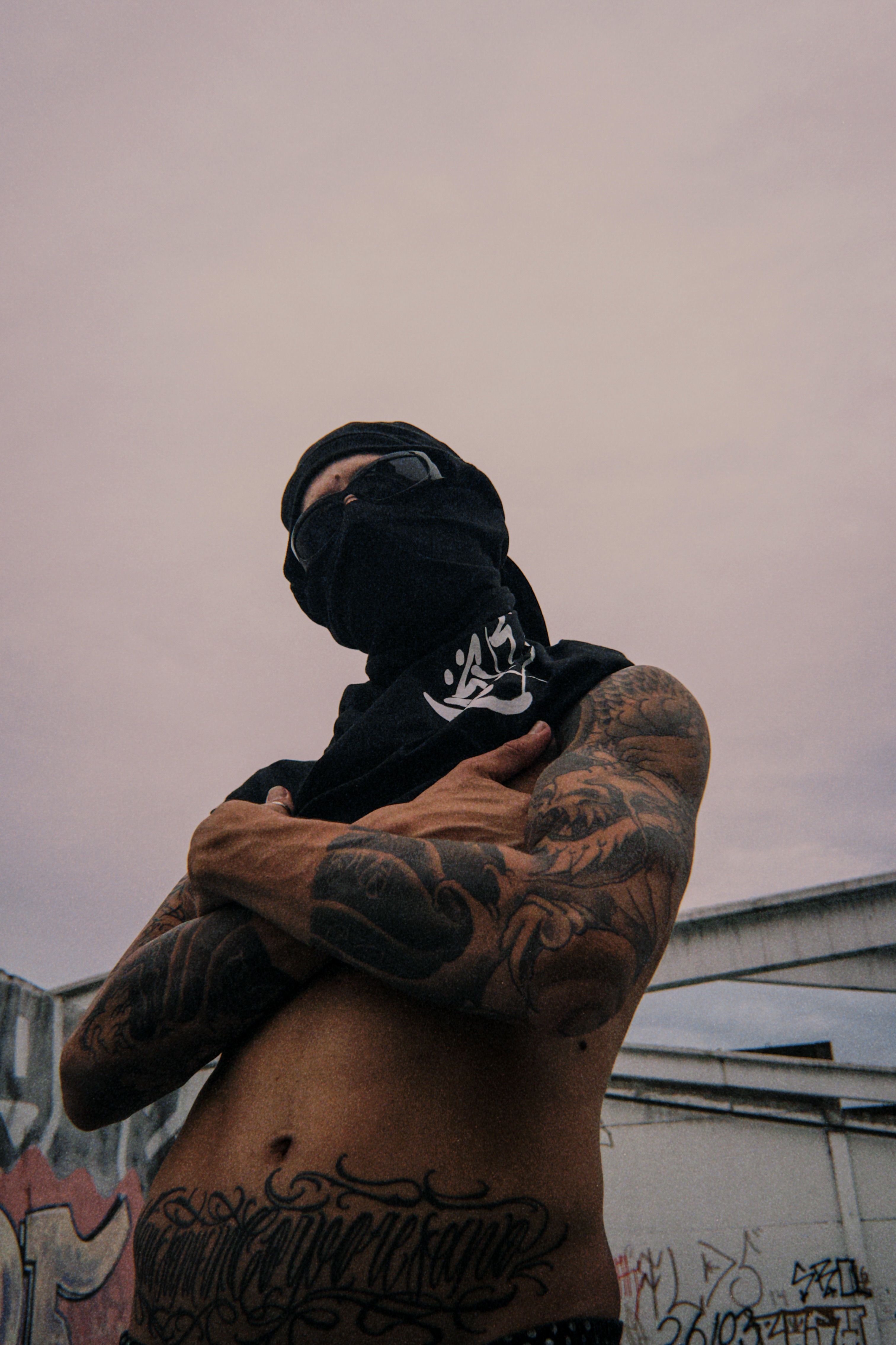 A tattooed person with face covered poses confidently outdoors in an urban area.