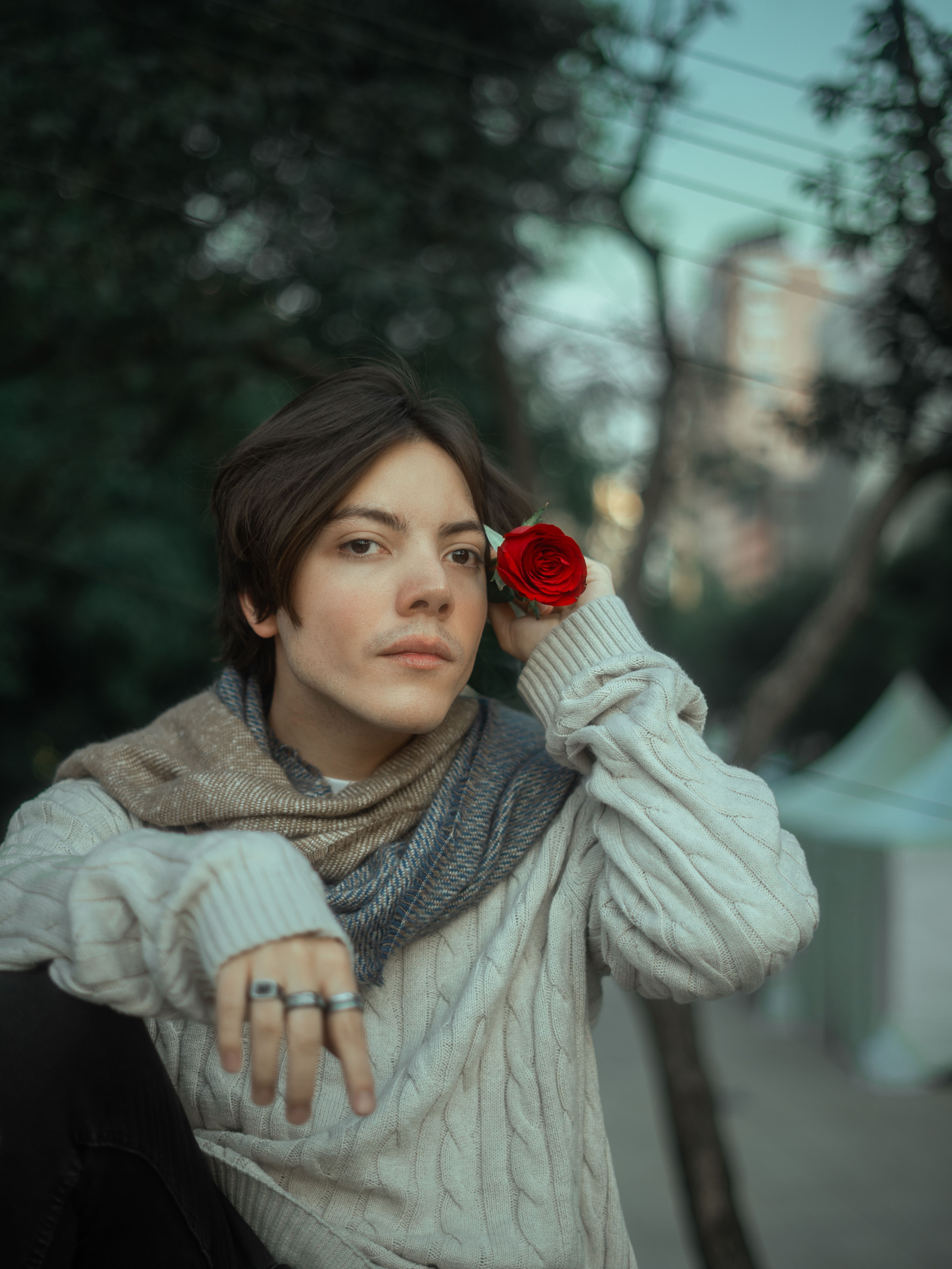 A young man holding a red rose poses outdoors in an artistic, romantic portrait.