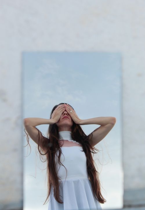 Free Woman in white dress covering eyes, standing outdoors with sky reflection. Stock Photo