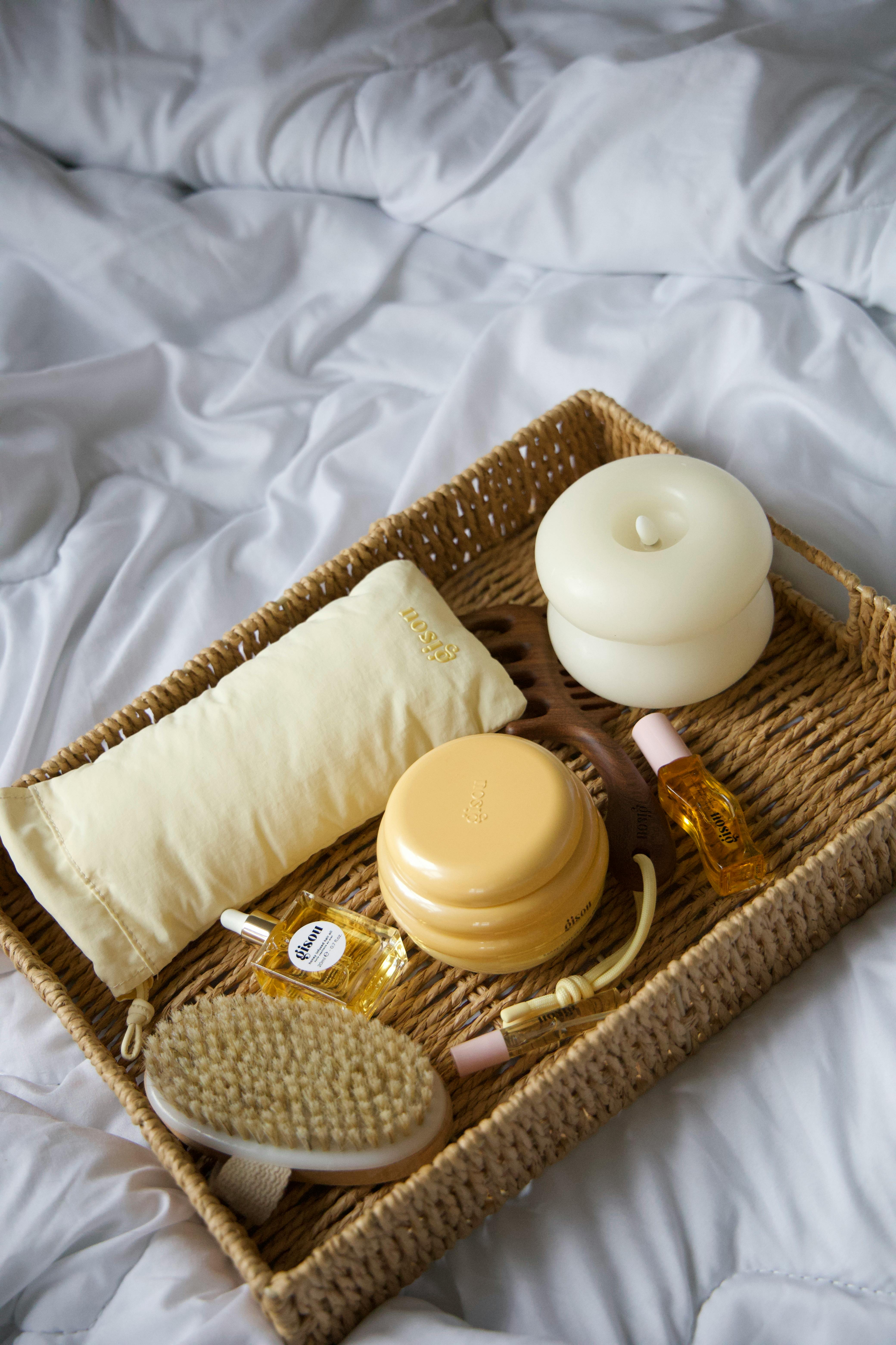 A cozy spa basket filled with massage tools, oils, and a cushion on a bed.