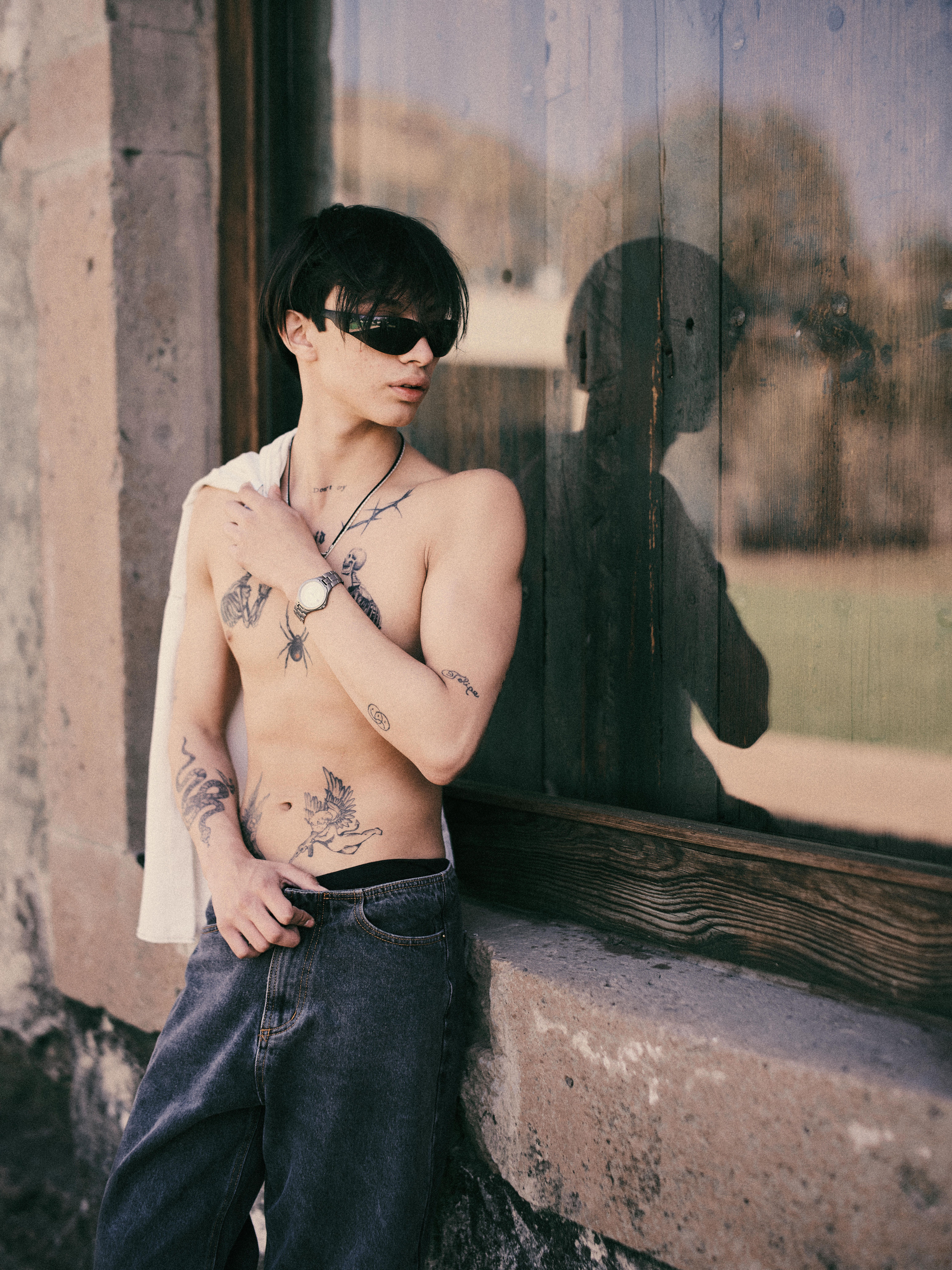Stylish tattooed man leaning against a stone wall, wearing sunglasses, and exuding confidence.