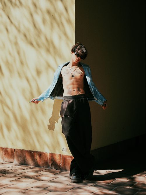 Free Young man in stylish attire poses against a sunlit wall, capturing a modern fashion vibe. Stock Photo