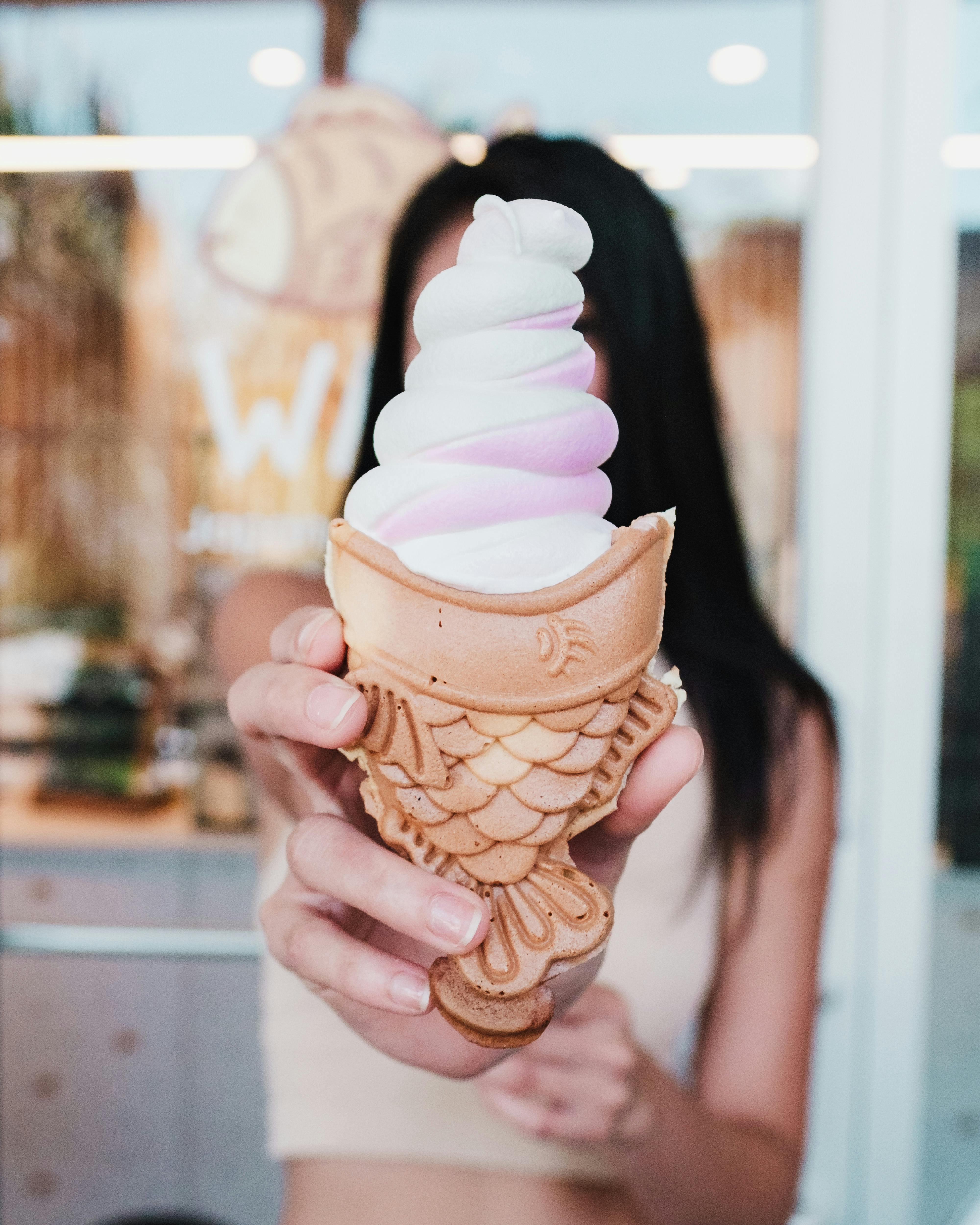 Close-up of a hand holding a taiyaki ice cream cone with pastel swirls, outdoors.
