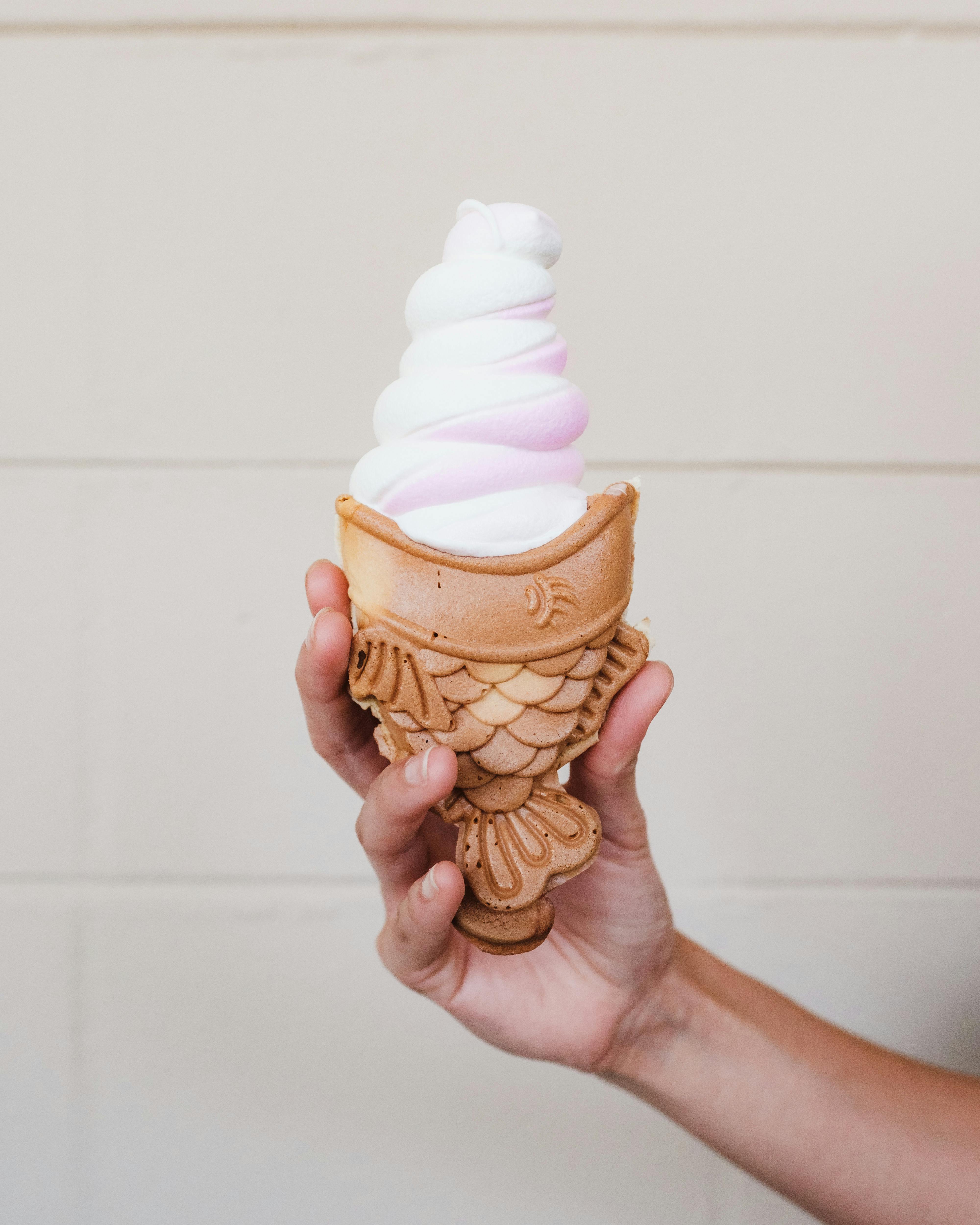 Hand holding a fish-shaped waffle cone filled with vanilla and strawberry swirl ice cream.