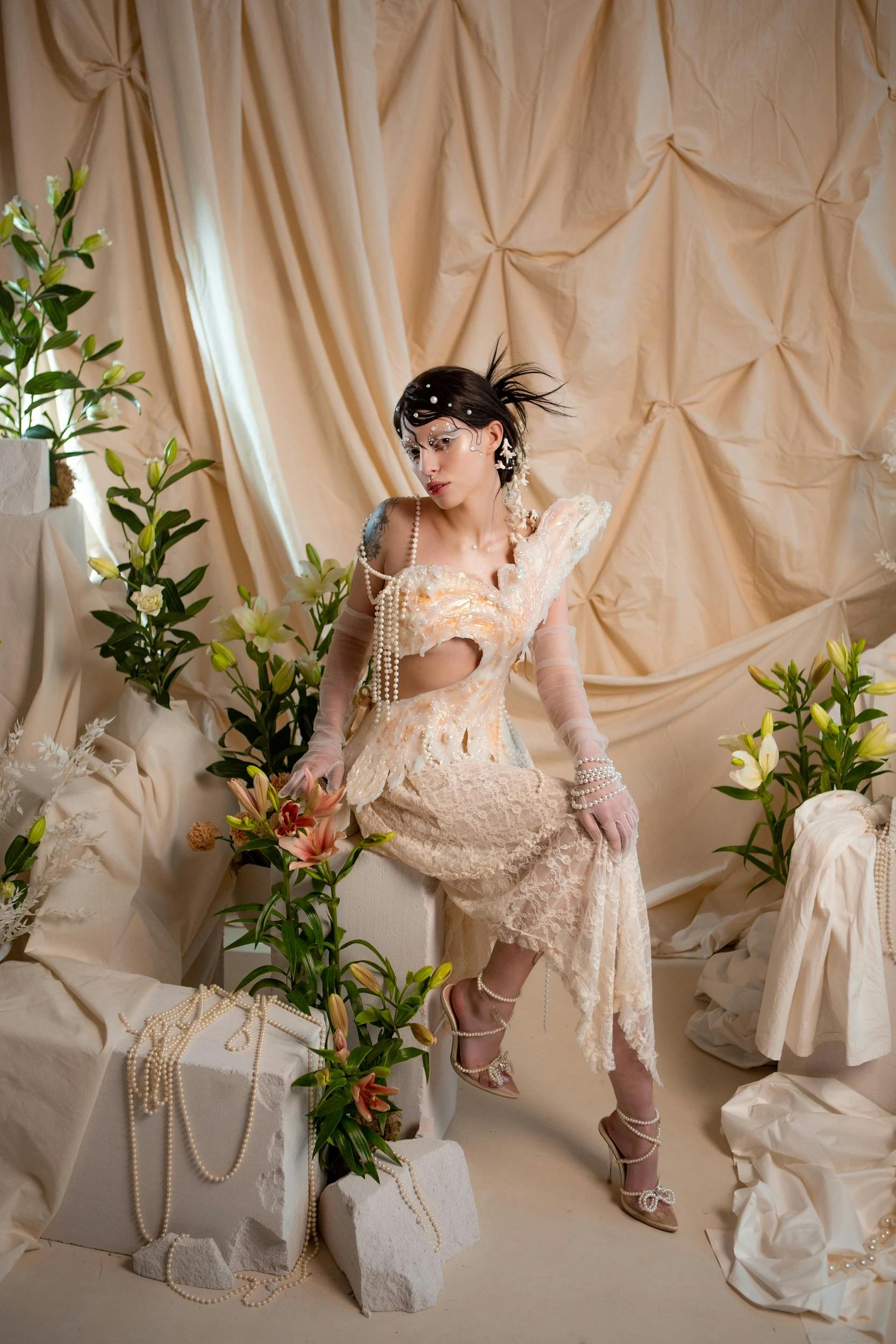 Fashionable woman in vintage attire surrounded by lilies and draped fabric.