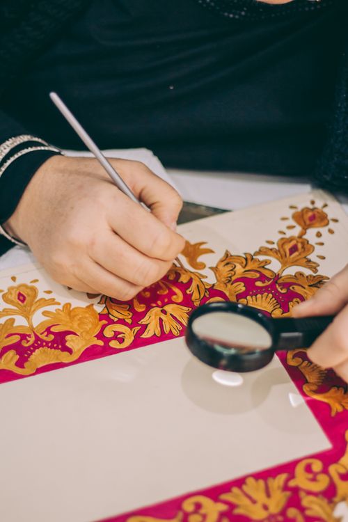 Free Artisan meticulously painting floral patterns on fabric with magnifying glass for precision. Stock Photo