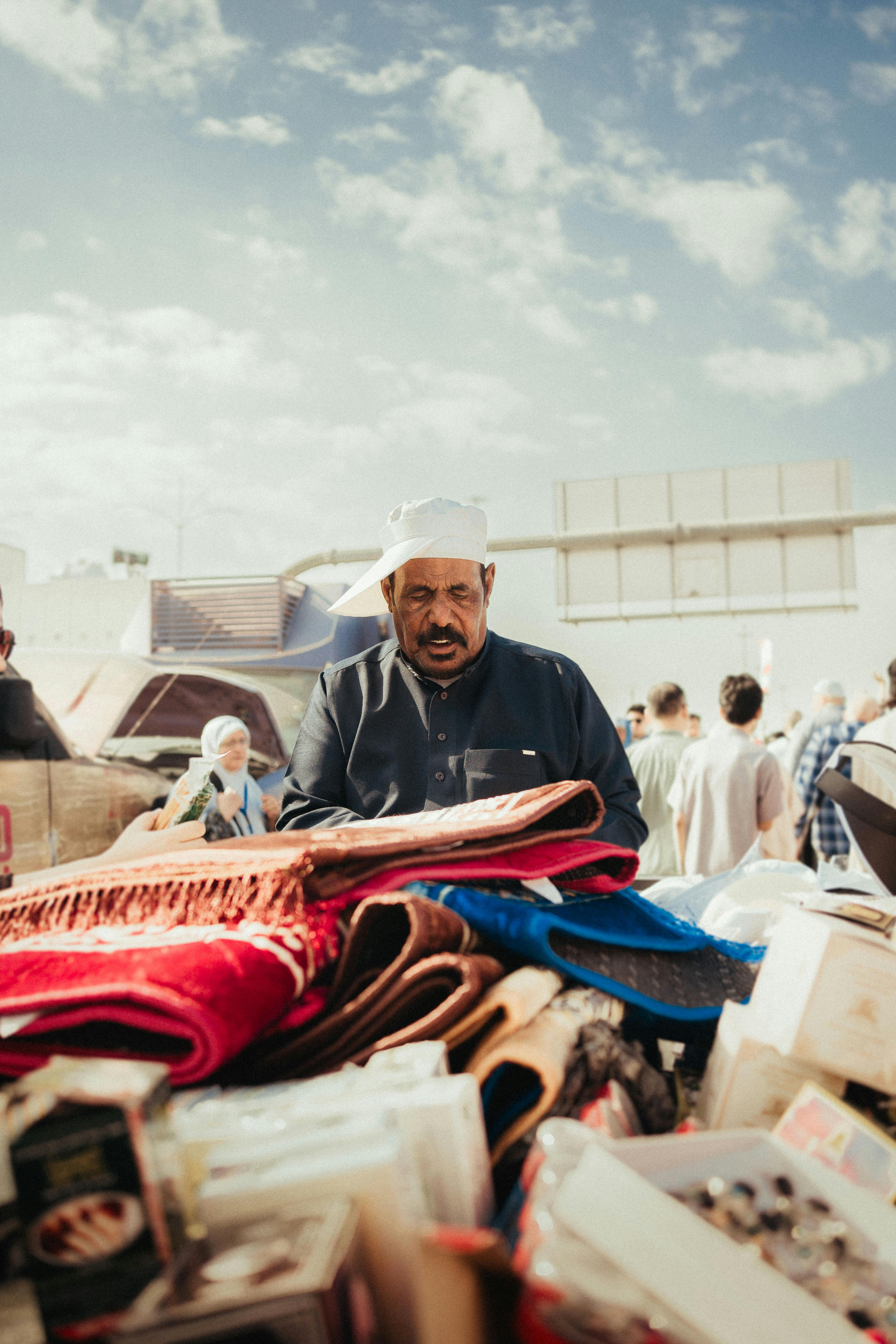 Man at Middle Eastern market stall selling textiles. Sunny day with crowd in background.