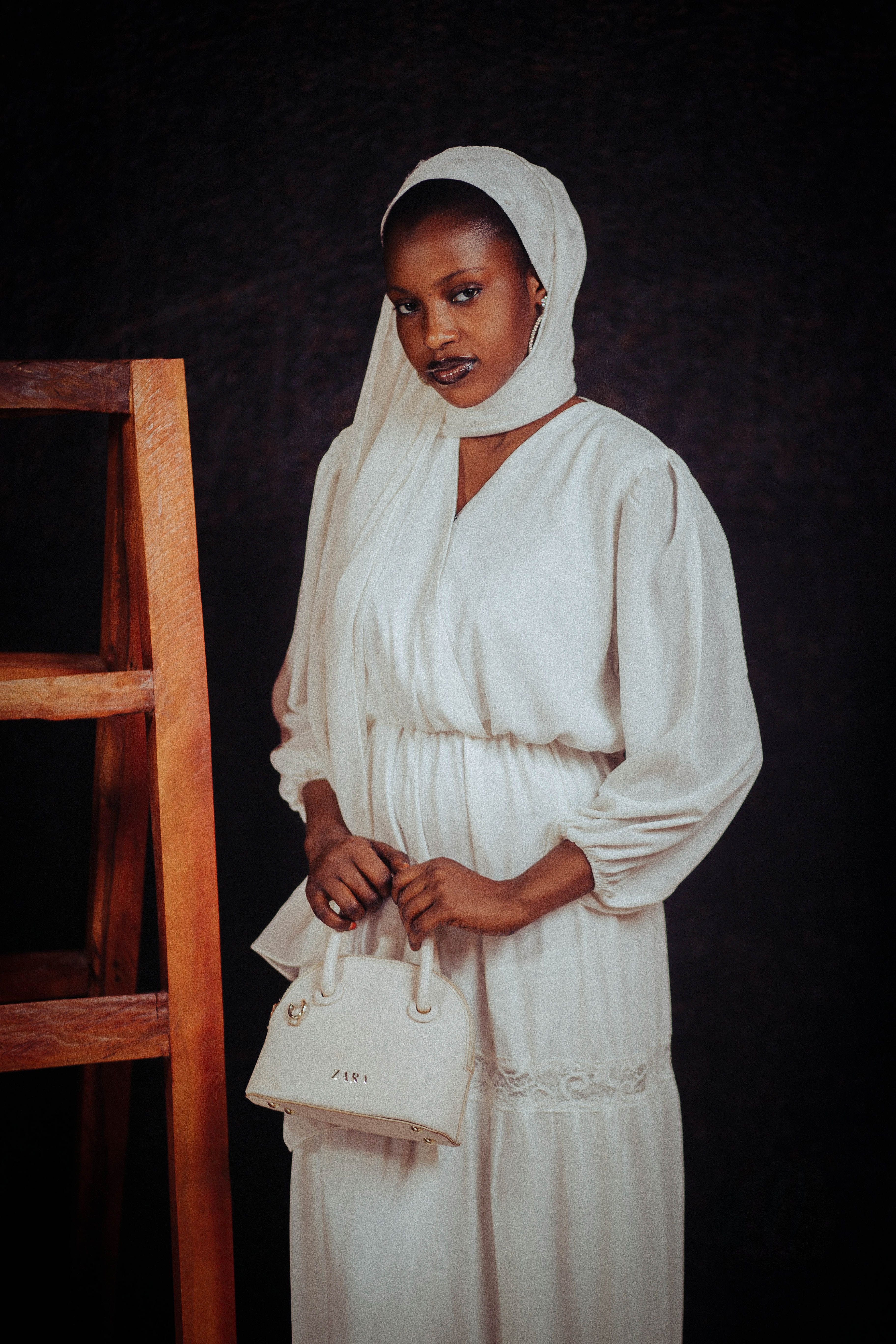 Stylish woman in white dress with headscarf stands confidently by a wooden ladder.