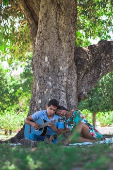 一对年轻夫妇在阳光明媚的户外，亲密地坐在树下弹着原声吉他