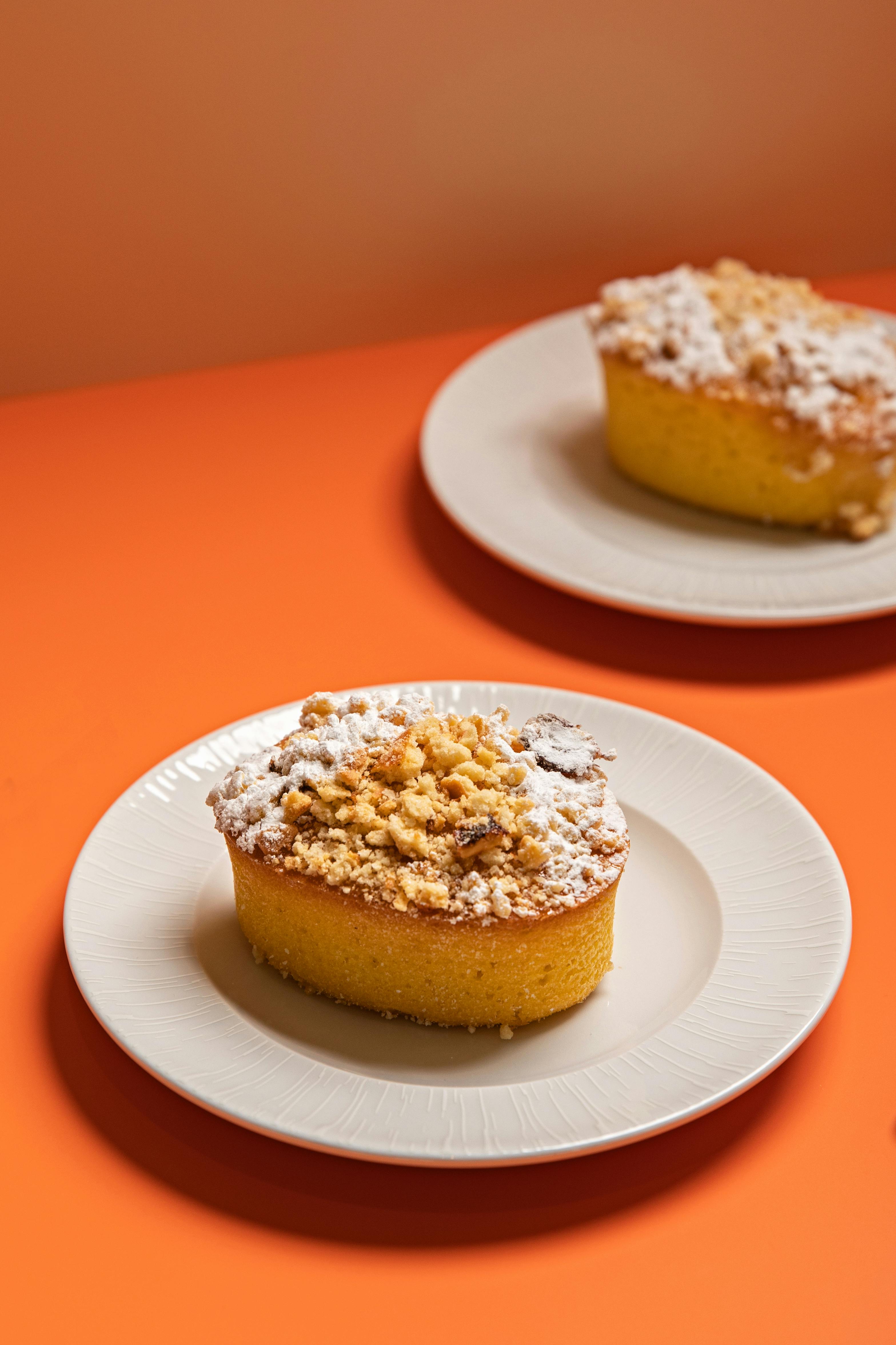 Two crumb cakes with powdered sugar on white plates against an orange background.