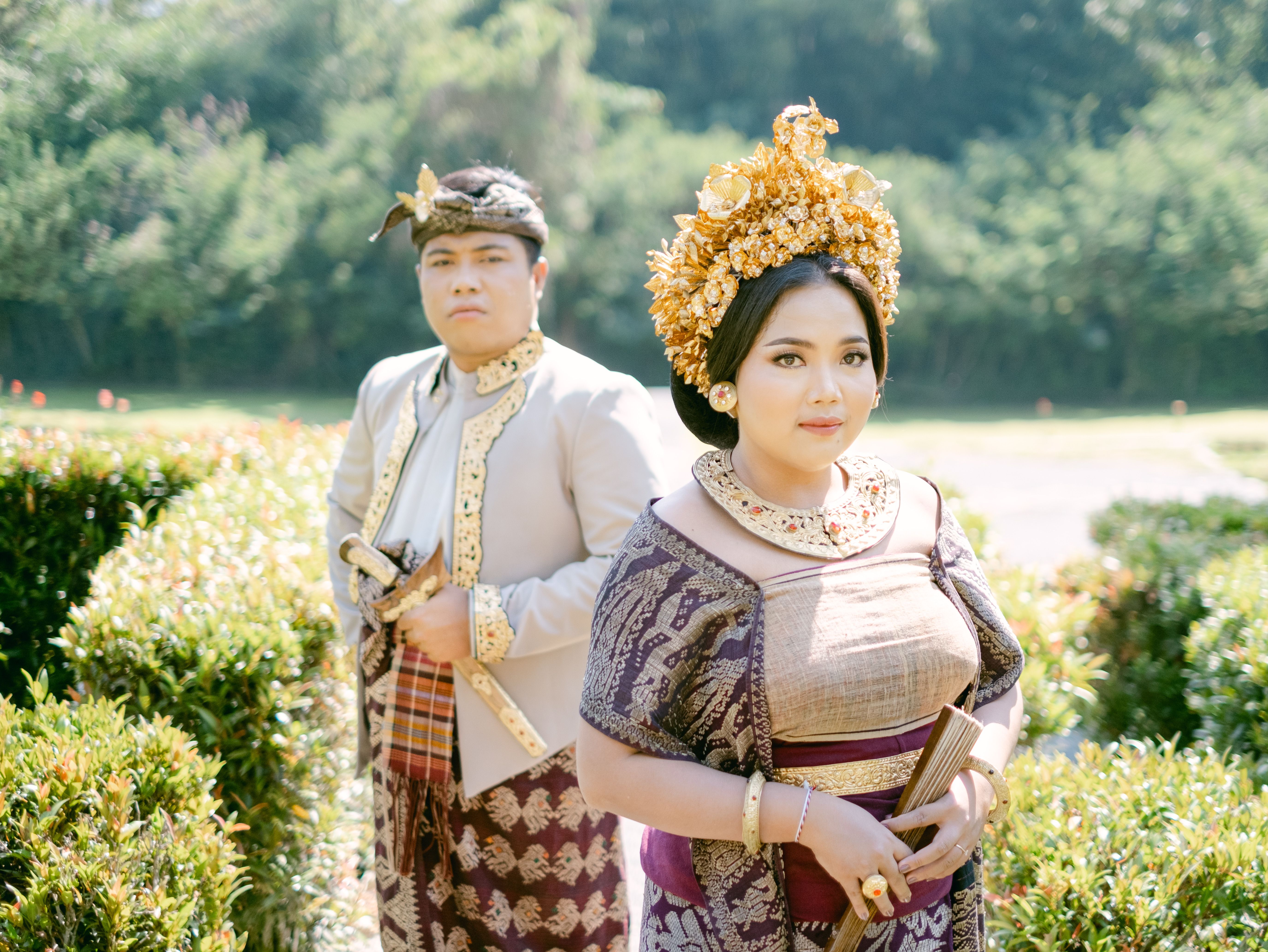 A couple dressed in traditional Balinese attire posing in a sunny garden setting.