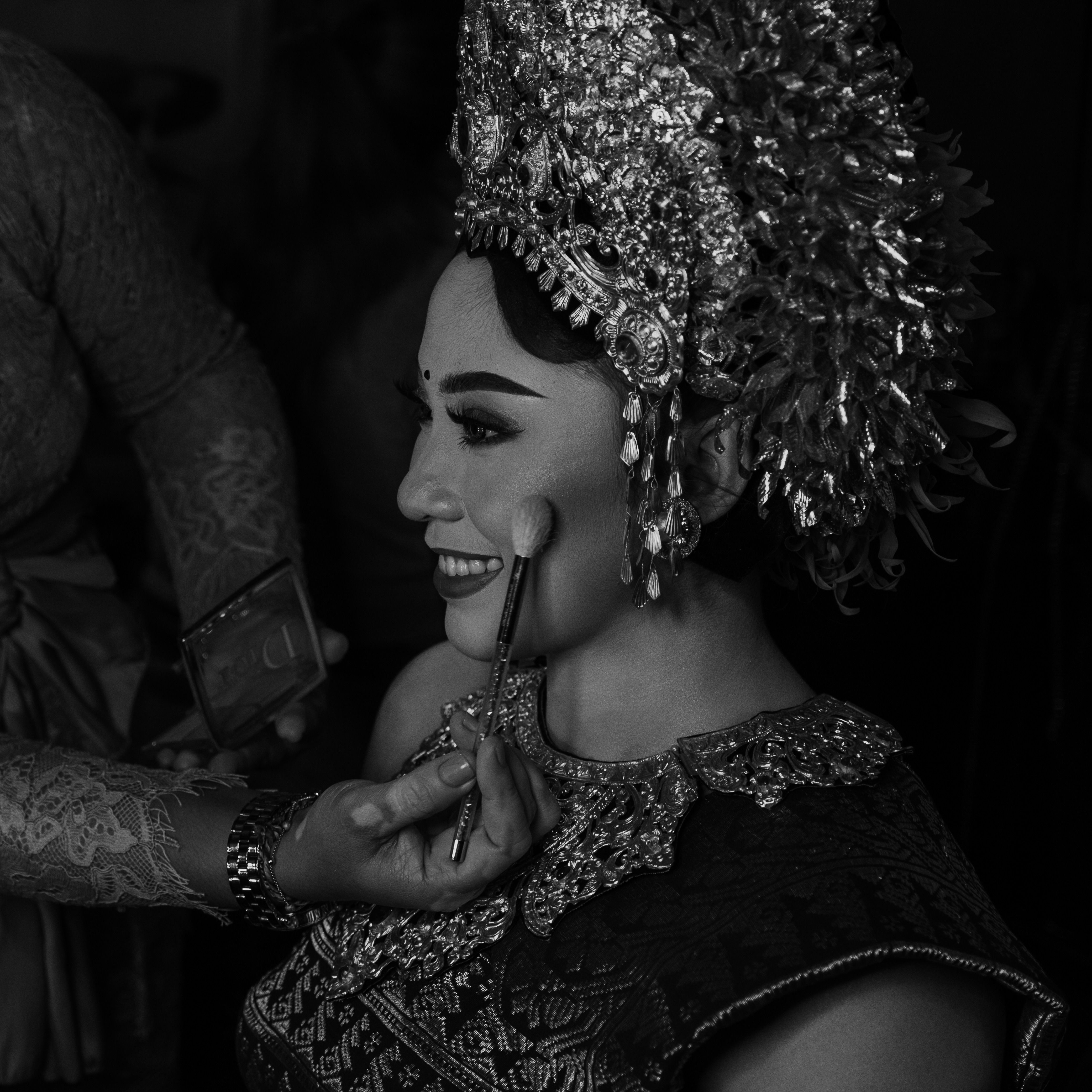 Black and white photograph of a bride in traditional attire receiving makeup touch-ups.