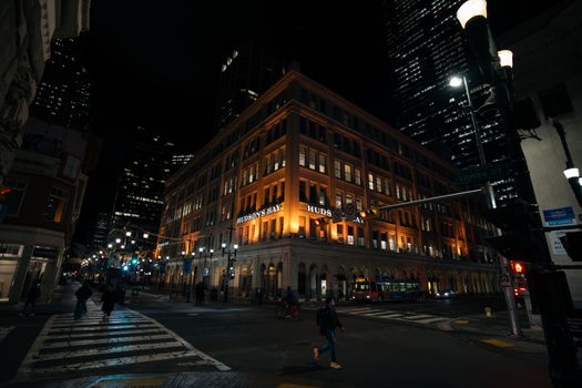 Illuminated Hudson's Bay building at night in bustling urban setting with pedestrians.