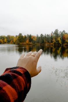 一只手伸向宁静的湖泊，周围秋树环绕，倒映着秋日的色彩