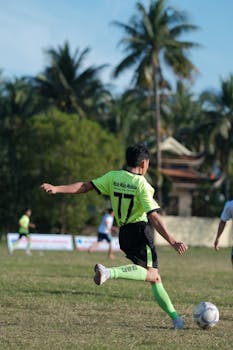 A soccer player kicks the ball during a match on a sunny field with palm trees.
