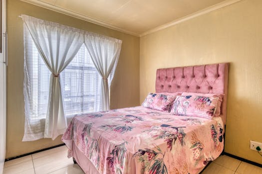 A cozy bedroom featuring pink floral bedding and natural light through sheer curtains.
