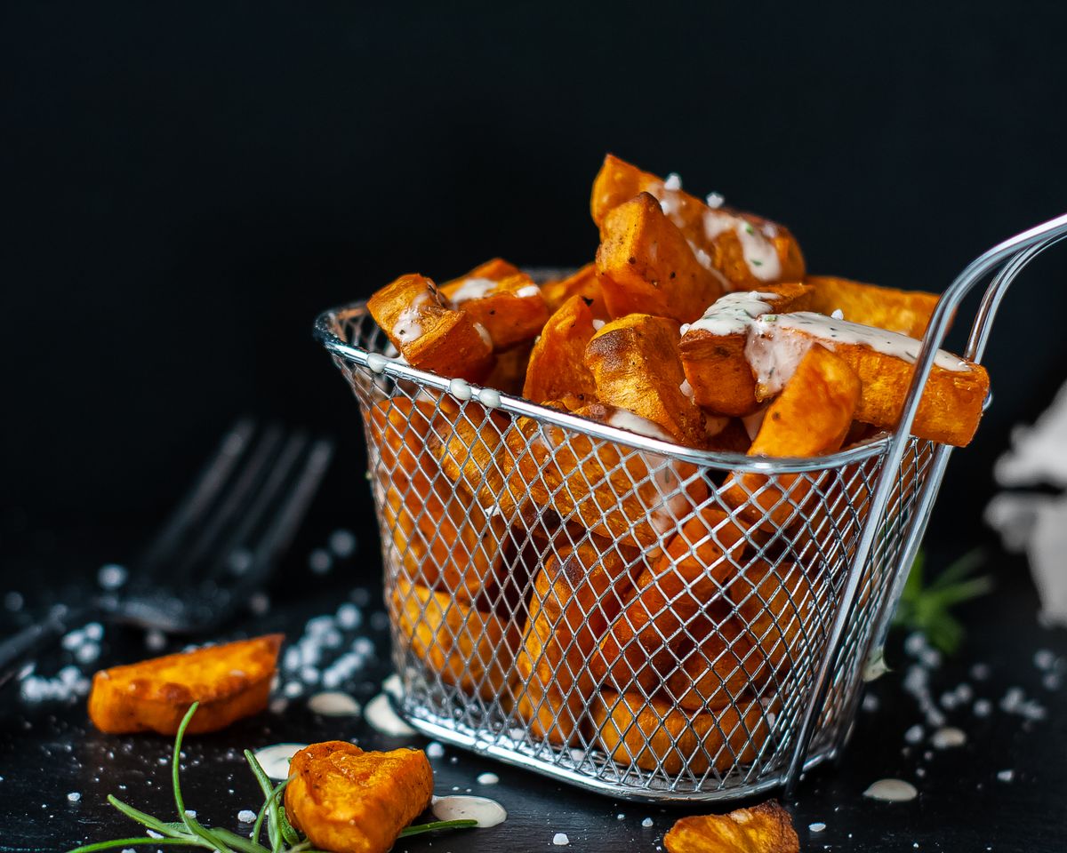 Wire fry basket filled with crispy sweet potato chunks drizzled with creamy sauce on dark slate