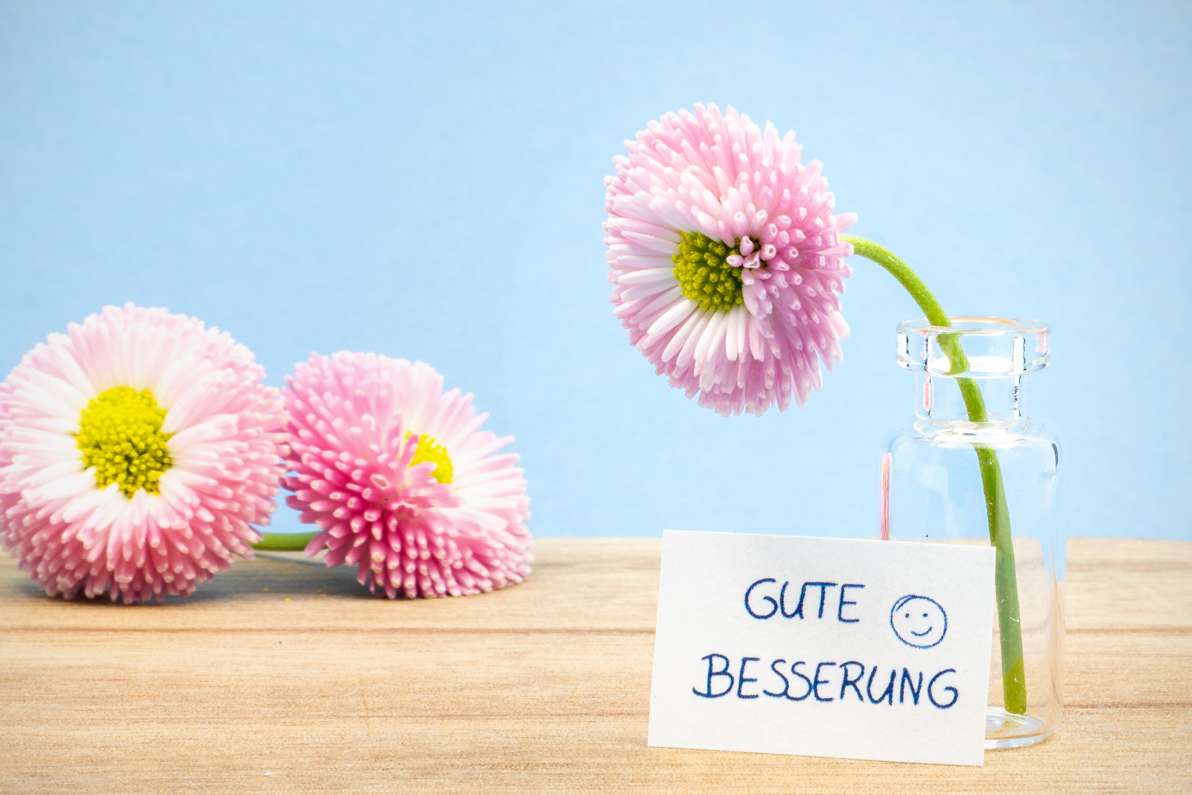 Pink daisies in a glass vase with a 'Gute Besserung' card on wooden table.