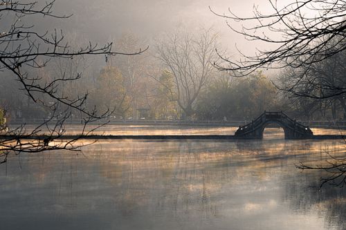 Free A tranquil scene of a misty pond and arch bridge at dawn, surrounded by bare trees. Stock Photo