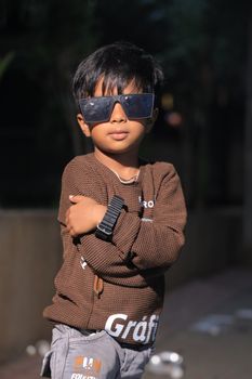 A young boy poses confidently wearing stylish sunglasses and a brown sweater.