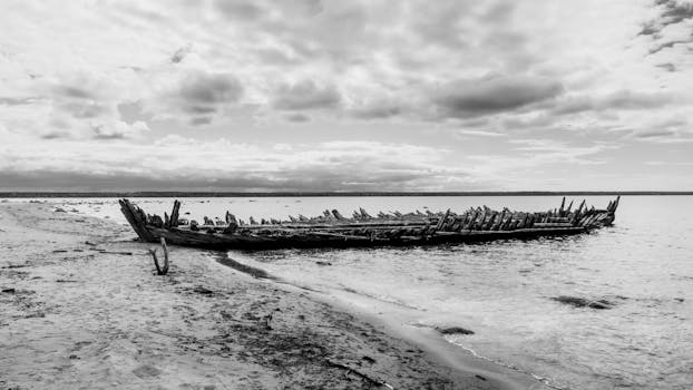 A dramatic black and white photo of an old shipwreck on a deserted beach with a cloudy sky.