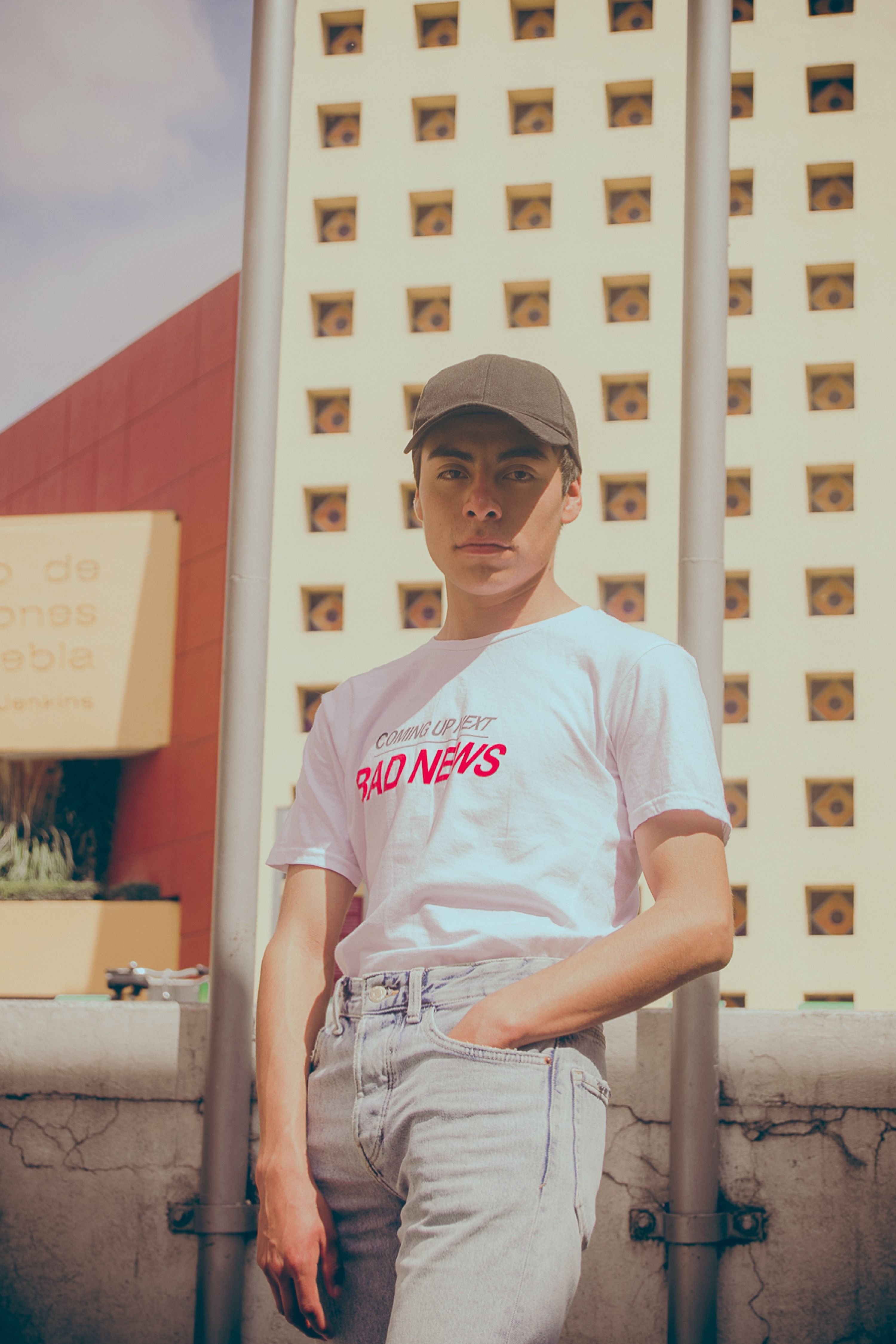 A young man wearing a cap and t-shirt poses in front of an urban architectural backdrop.